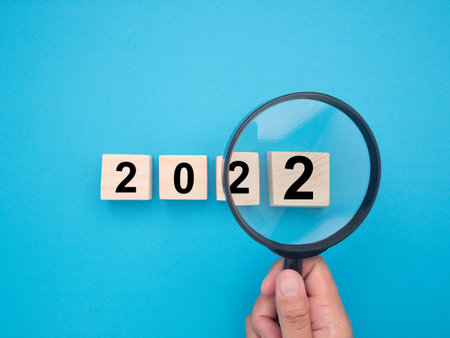 Hand of holding a magnifying glass looking to ideas for the 2022 year. Close-up photo. Top view of wooden blocks with words 2022. Space for text.の写真素材