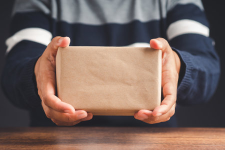 Man in sweater holding a brown box on a wooden table. A box wrapped in brown paper. Close-up photo. Concept of Christmas, New Year, Anniversary, Happy birthday, etcの写真素材