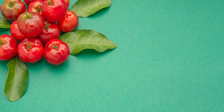 Pile of ripe red acerola cherries and green leaves isolated on a green background. High vitamin C and antioxidant fruits. Top view. Space for text. Close-up photo. Healthy fruits concept.の写真素材