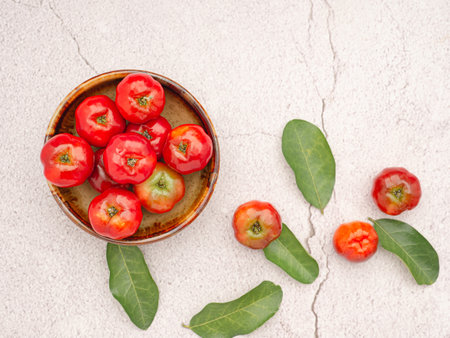 Healthy fruit concept. Ripe red acerola cherries fruit in a ceramic bowl and green leaves on a cement floor. High vitamin C and antioxidant fruits. Top view. Space for textの写真素材
