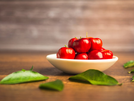 Ripe red acerola cherries fruit in a ceramic bowl and green leaves on a wooden table with a wooden wall background. High vitamin C and antioxidant fruits. Space for text. Concept of healthy fruitsの写真素材