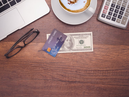 Working desk table concept. Top view of a mockup blue credit card over on dollar banknotes, a laptop, calculator, and a coffee cup on a wooden table in the office. Flat lay, space for textの写真素材