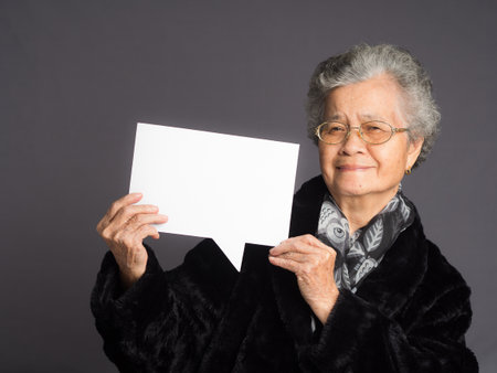 Concept of a speech bubble. Happy an elderly Asian woman in a sweater with short gray hair holding a blank paper and looking at the camera with a smile while standing on a gray backgroundの写真素材