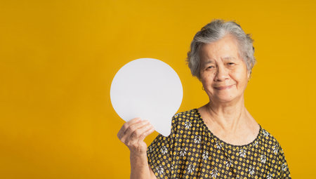 Concept of a speech bubble. Happy senior woman with short gray hair holding a blank paper and looking at the camera with a smile while standing on a yellow background in the studioの写真素材