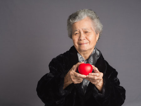 An elderly Asian woman in a sweater with short gray hair holding a red heart shape and looking at the camera with a smile while standing on a gray background. Aged people and healthcare conceptの写真素材