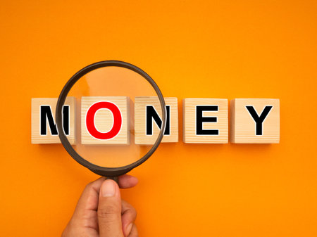 Money concept. Top view of hand holding a magnifying glass reading words MONEY on wooden cubes on a yellow background. Close-up photoの写真素材