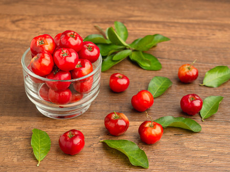 Ripe red acerola cherries in glass and green leaves scattered on a wooden table. High vitamin C and antioxidant fruits. Space for text. Close-up. Healthy fruits conceptの写真素材