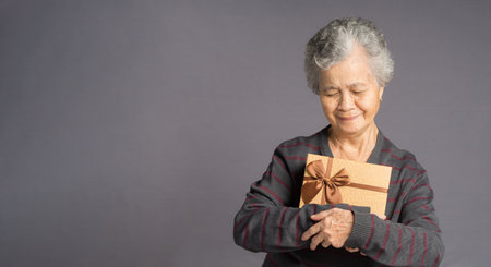 Portrait of an elderly Asian woman with short gray hair wearing a sweater holding and looking at a brown gift box with a smile while standing over a gray background. Space for textの写真素材