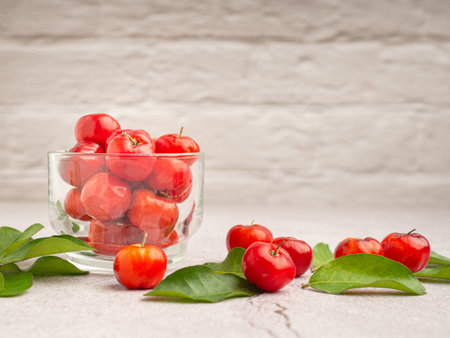 Ripe red acerola cherries in glass and green leaves scattered on a cement floor with a white brick background. High vitamin C and antioxidant fruits. Space for text. Healthy fruits conceptの写真素材