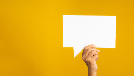 A speech bubble concept. Hand holding of an empty white speech balloon against a yellow background. Space for text. Close-up photoの写真素材