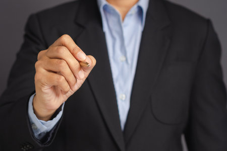 Businessman in suit holding pen while standing over gray background with space for text. Ready to write something. Focus on the pen. Close-up photoの写真素材