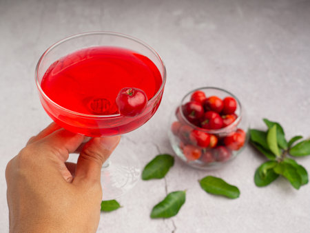 Hand holding a wine glass with cherry juice. Ripe red acerola cherries in glass and green leaves on a cement floor. High vitamin C and antioxidant fruit. Close-up photoの写真素材