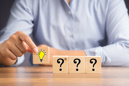 Question mark and creativity concept. Hand holding a wooden cube with a drawn light bulb and a row of cubes with question marks on the wooden tableの写真素材