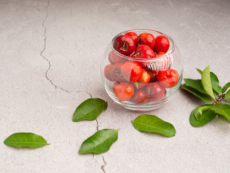 Overhead view of ripe red acerola cherries in a glass bowl and green leaves scattered on a cement floor. High vitamin C and antioxidant fruits. Space for text. Close-up. Healthy fruits conceptの写真素材