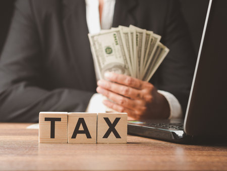 Tax payment and Calculation tax return concept. Close-up of wooden cubes with letters "TAX" with a businessman in a suit holding US dollars banknote blurred backgroundの写真素材