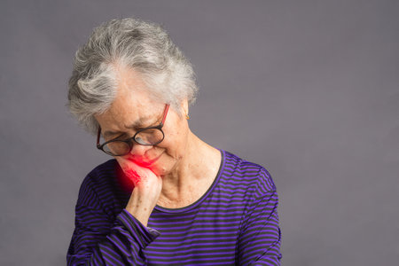 An elderly Asian woman with short gray hair wearing glasses has a toothache while standing on gray...