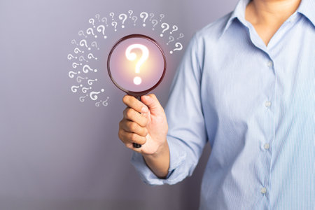 Question mark concept. A businessman is holding a magnifying glass looking at a question mark symbol while standing on a gray background.の写真素材