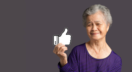 Service rating, satisfaction concept. Portrait of an elderly Asian woman holding a thumbs up a symbol and looking at the camera with a smile while standing on a gray background. Space for textの写真素材