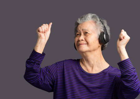 Cheerful elderly Asian woman with short gray hair wearing wireless headphones to listen to a...