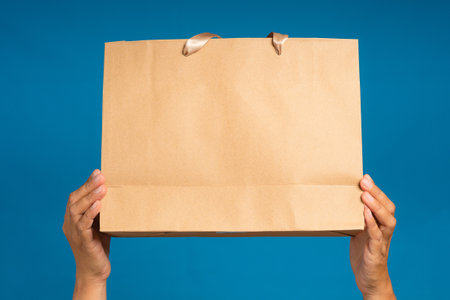 Hands holding a brown paper bag against on blue background. Packaging template mockup. Delivery...