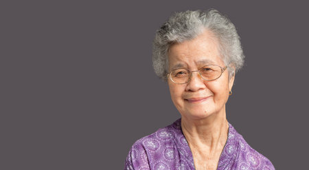 Cheerful an elderly Asian woman with short gray hair looking at the camera with a smile while standing on gray background in the studio. Space for text. Aged people and relaxation conceptの写真素材