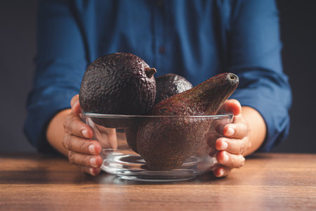 Close-up of hand holding a glass bowl with fresh avocados over a wooden table. Healthy fruit and...