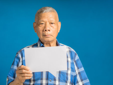 A speech bubble concept. Senior man holding a blank paper speech bubble and looking at the camera while standing on a blue background in the studio. Space for textの写真素材