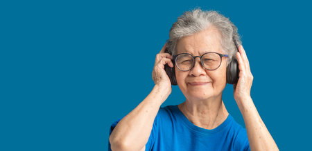 Senior woman wearing wireless headphones to listen to a favorite song and looking at the camera with a smile while standing on a blue background. Space for text. Aged people and relaxation conceptの写真素材