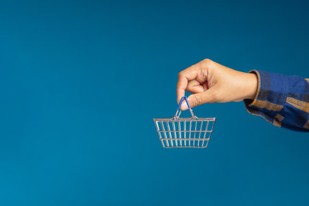 Business and e-commerce concept. Close-up of hand holding an empty mini shopping cart over a blue background. Side view. Space for textの写真素材
