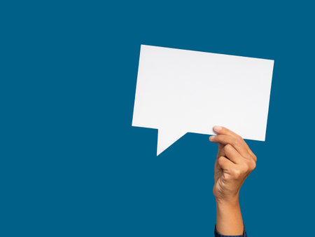 A speech bubble concept. Hand holding of an empty white speech bubble against a blue background. Space for text. Close-up photoの写真素材