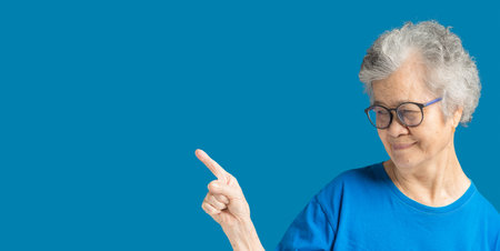 Portrait of an elderly Asian woman with short gray hair wearing glasses pointing the finger and a smile while standing over a blue background. Aged people and relaxation conceptの写真素材