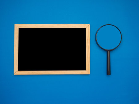A mini blank blackboard with a wooden border and magnifying glass over a blue background. Space for text. Top viewの写真素材