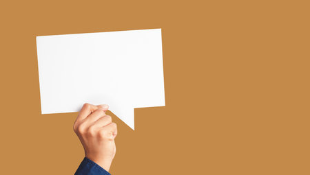 Close-up of hand holding a white speech bubble against a light brown background in the studio....