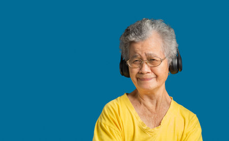 Senior woman with short gray hair wearing wireless headphones to listen to a favorite song and arms crossed with a smile while standing on a blue background. Aged people and relaxation conceptの写真素材