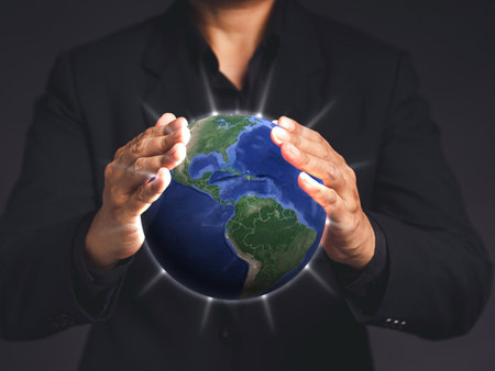 A businessman in a black suit hands protecting the earth on a virtual screen while standing on a gray background.の写真素材