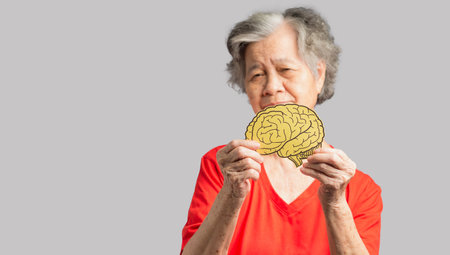 Portrait of a senior woman holding a brain shape made from yellow paper against a gray background....