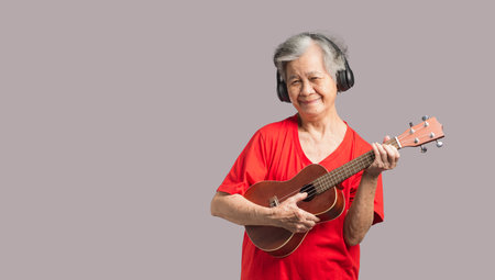 Portrait of an elderly Asian woman with short gray hair playing the ukulele with a smile while standing on a gray background. Enjoy life after retiring. Concept of aged people and relaxationの写真素材