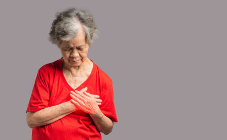 An elderly Asian woman in a red casual has sore left chest pain while standing over a gray background in the studio. Close-up photo. Space for text. Concept of aged people, medical and healthcareの写真素材
