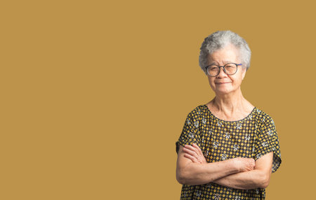 Portrait of an elderly Asian woman with short gray hair looking at the camera with a smile while standing arms crossed on a brown background in the studio. Aged people and relaxation conceptの写真素材
