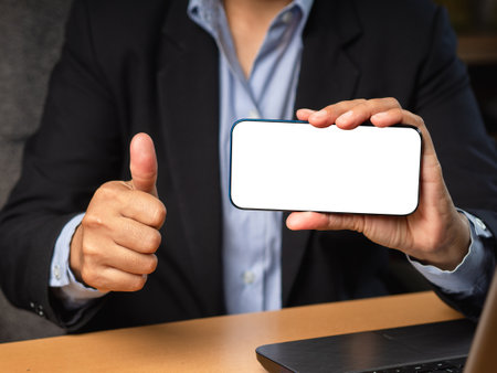 Hand holding a horizontal mobile phone with a blank white screen and showing thumbs up while sitting in the office. Customer feedback, service evaluation, experience, and satisfaction conceptの写真素材