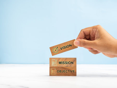 Hand holding a wooden cube with the letters Vision, Mission, and objective on a marble table against a blue background. Business and strategy conceptの写真素材