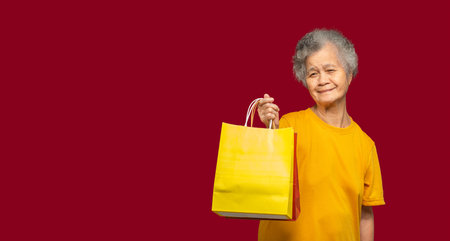 A portrait of a senior woman is holding yellow and red paper bags with a handle looking at the camera with a smile while standing on a red background. Aged people and shopping conceptの写真素材