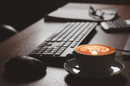 Close-up of a white coffee cup with shape heart latte art with a keyboard and mouse at the office desk. Relax, beverage, and workplace conceptの写真素材