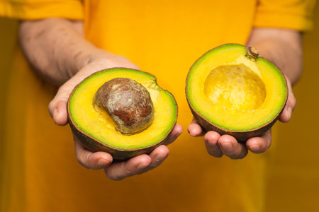 Close-up of hands holding a fresh avocado while standing on a yellow background. Healthy fruit and...