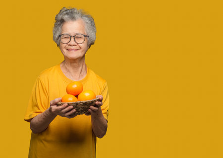 Senior woman's hands holding a basket with fresh oranges while standing on a yellow background.の写真素材