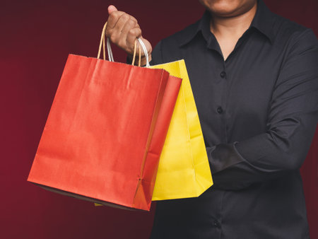 Close-up of hand holding a yellow and red paper bag with a handle while standing on a red background. Packaging template mockup. Delivery service, shopping, and advertising area conceptの写真素材