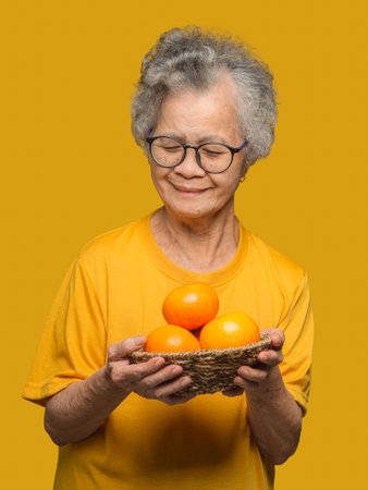 Senior woman's hands holding a basket with fresh oranges while standing on a yellow background. Healthy fruit conceptの写真素材