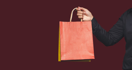 Close-up of hand holding a red paper bag with a handle while standing on a red background. Packaging...