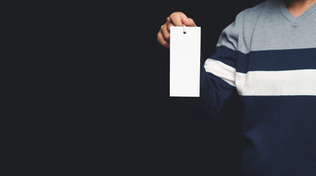 Hand holding a blank white label or price tag while standing on a black background. Space for text. Close-up photo. Shopping and price tag conceptの写真素材