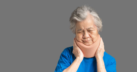 Portrait of a senior woman wearing a soft neck collar because suffering neck pain and neck tilt while standing on a gray background. Medical and healthcare conceptの写真素材